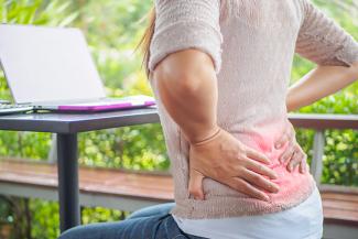 A woman in a beige top holding the small of her back which is marked red to indicate back pain looking at a laptop on the table in front of her