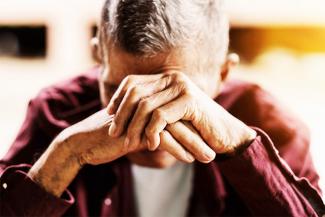 Image showing an elderly person in a red shirt over a white T-shirt reacting to verbal abuse and neglect by placing his head over his hands