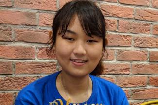 A young woman with bangs and wearing a blue tshirt in front of a red brick wall