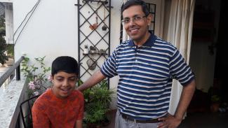 A young autistic boy on the left in a red shirt with his father in a striped blue and white shirt in a home balcony