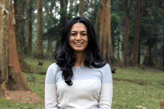 A young lady with dark hair in a white full sleeved dress in a green surrounding with trees in the background