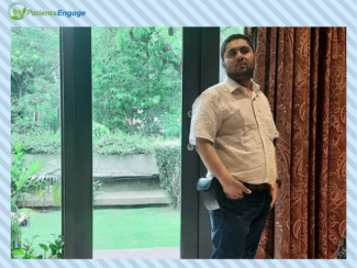 Mohit Gupta in a white shirt and trousers standing in front of a glass window covered partially by a curtain. Behind the glass window is a green garden