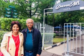 Rita Banik on the left in a red dress and beige jacket with her husband on the right with a blue shirt and black jacket standing by the gate of a building and a garden in the background