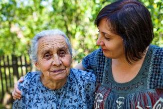 A young woman with short dark hair on the right supporting and looking at an elderly silver haired woman on the left