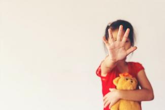 A child in a red tshirt holding a baby doll and stretching a hand out to block. The stretched hand covers the face