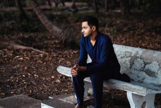 A man in a dark shirt and trousers sitting on a bench in front of a clearing of trees