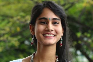 Image of Sunena Gupta, a black haired woman with alopecia smiling and facing the camera against a background of green foliage