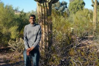 A young man in grey shirt and blue jeans with cactus and other green plants in the background