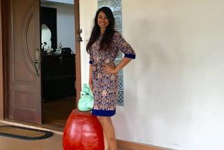 A young person in a printed blue and gold dress standing next to a red stool which has a green artifact on top. In the background a window panel and a half open door through which you can see a room inside