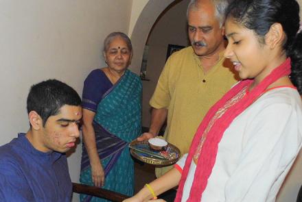 Image Description: Nivriti on the right in a white kurta and red dupatta tying a rakhi on her autistic brother Nishant's wrist. Nishant is sitting and wearing a blue kurta. Looking over both of them  is their father in a mustard kurta and grandmother in a blue sari