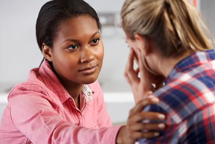 Image Description: Stock pic of a black haired woman on the left in a pink shirt supporting a young blond haired woman in a blue, red and white checked shirt on the right whose back is visible to us