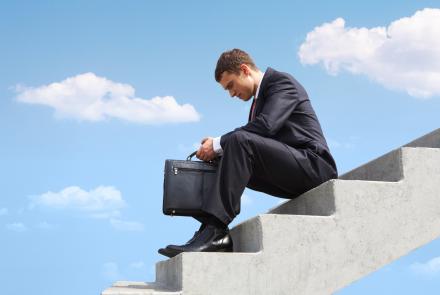 A man in a suit and with his work bag, sitting on steps, looking downwards and worried