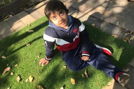 A young boy in navy blue pants, and blue, red and white jacket and red and white shoes sitting on the green grass and looking up at the camera