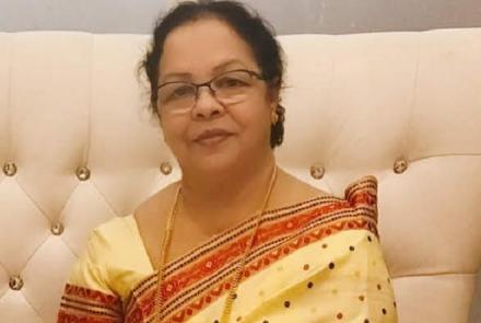 A dark haired woman with spectacles in a yellow and red sari and blouse sitting on a white sofa